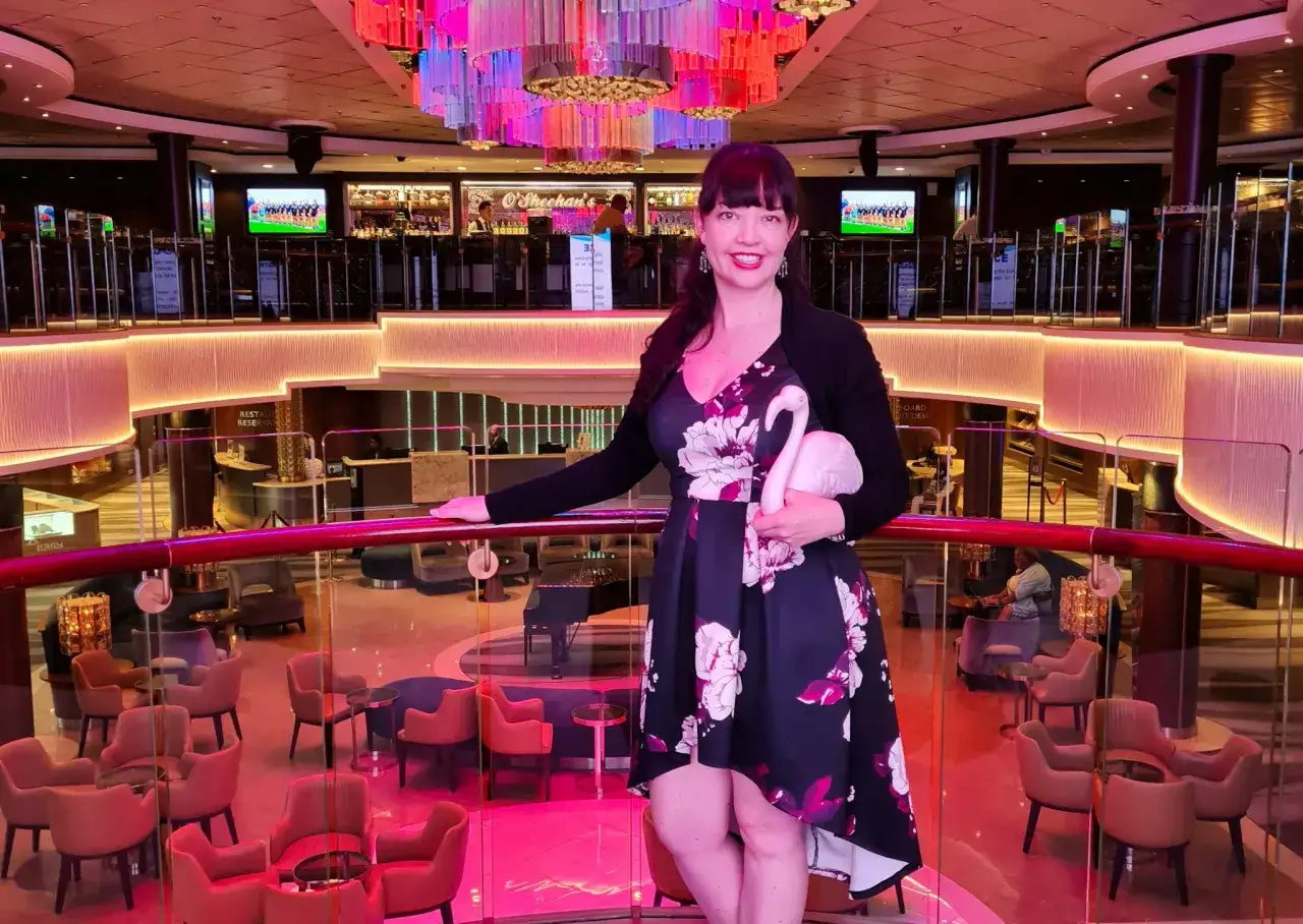 Donna sorridente con fenicottero rosa, felice di lavorare sulle navi da crociera. L'ambiente è un lounge elegante con luci colorate e arredi moderni.