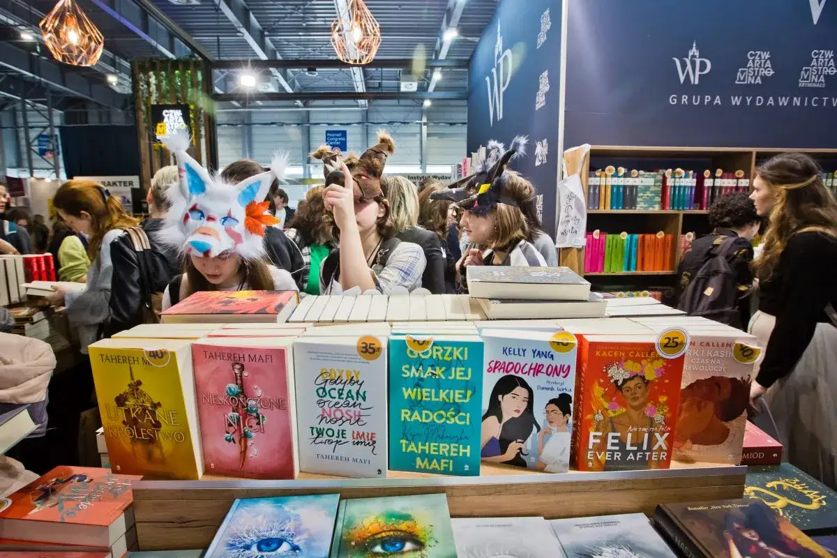 Targi książki: jak przygotować się do nich jako autor i czytelnik. Widać stoisko z książkami, ludzi w kostiumach i okładki bestsellerów.
