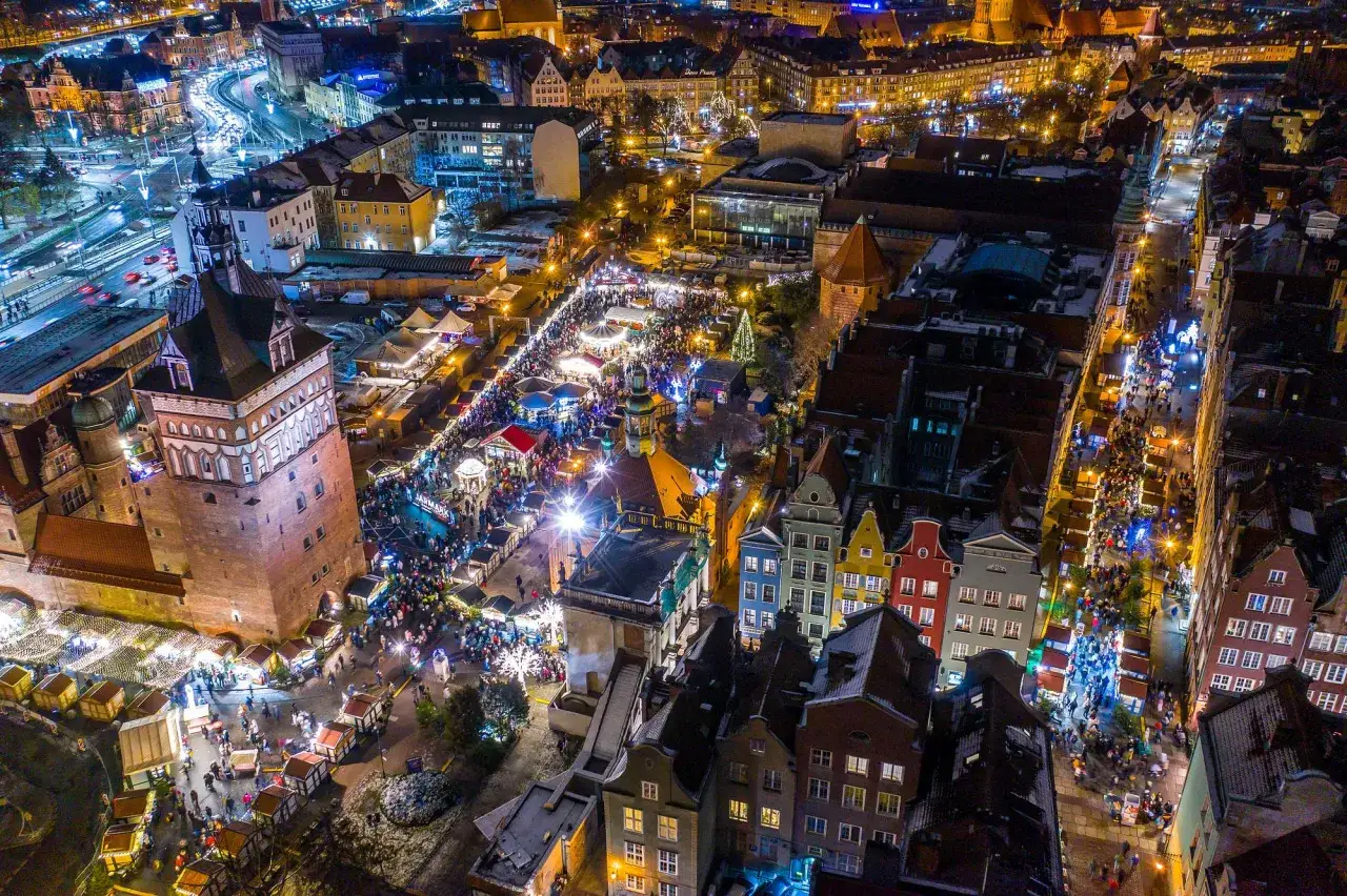 Nocny Gdańsk rozświetlony jarmarkiem świątecznym. Tłumy ludzi spacerują między straganami, podziwiając iluminacje i świąteczną atmosferę.