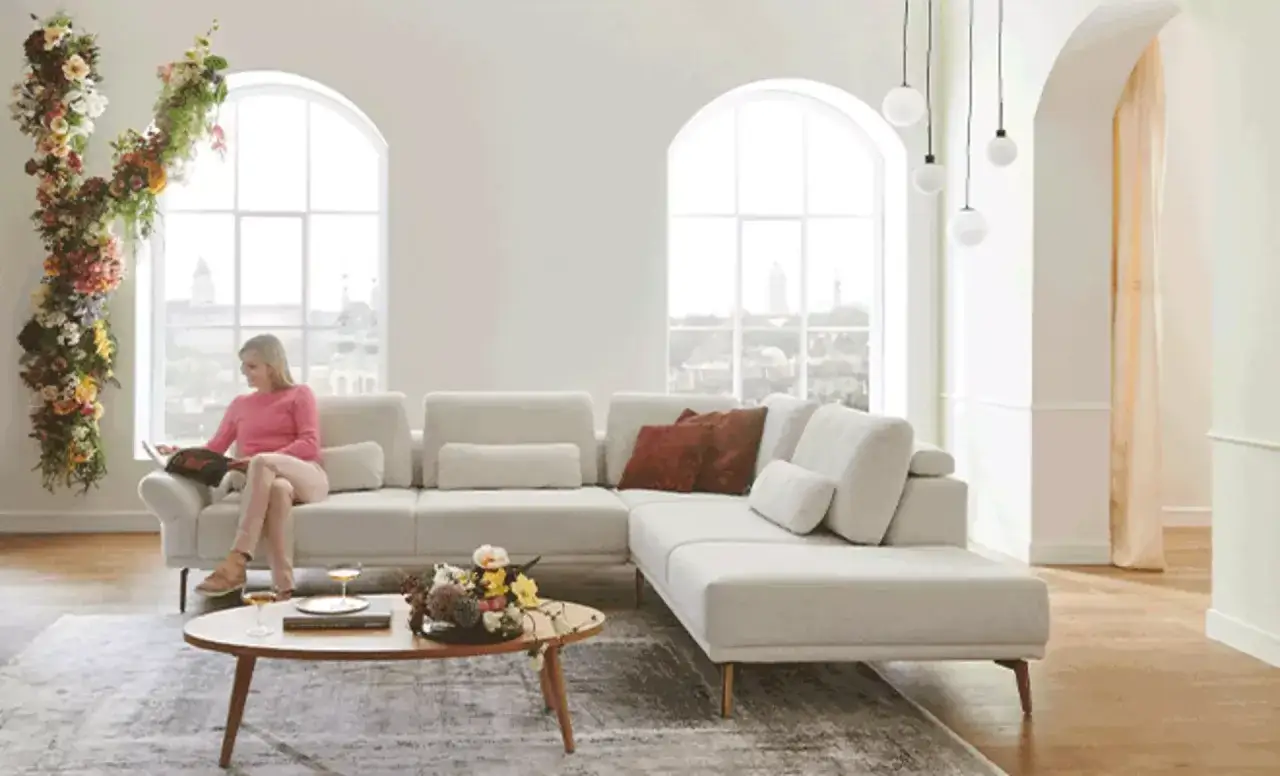 Frau liest auf einem beigen Sofa in einer hellen Wohnung. Blumenranken schmücken das Fenster. Perfekt, um die Wohnung einzurichten.