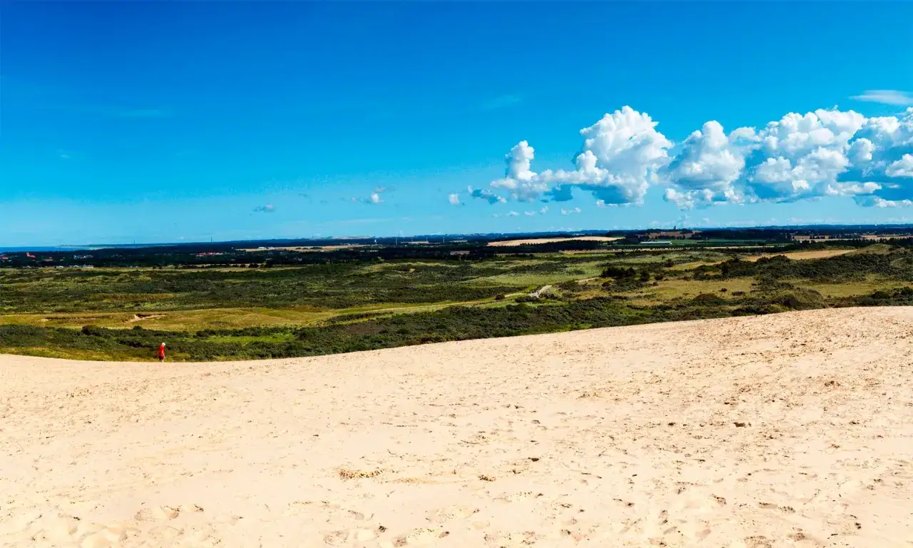 En person i r&ouml;d jacka vandrar p&aring; sanddyner i Danmark. Landskapet str&auml;cker sig mot en bl&aring; himmel med moln.