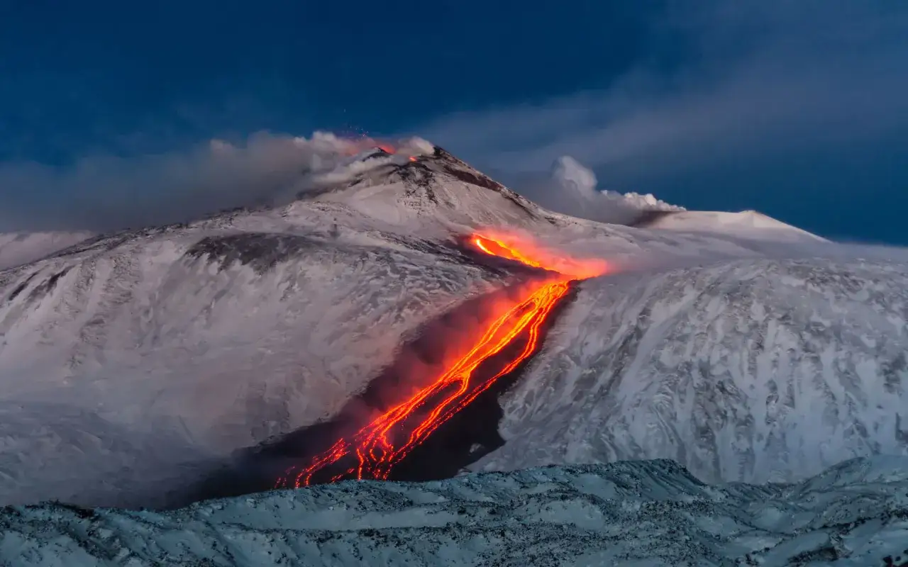 Sci Etna in eruzione, con lava incandescente che scorre su pendii innevati sotto un cielo blu scuro.