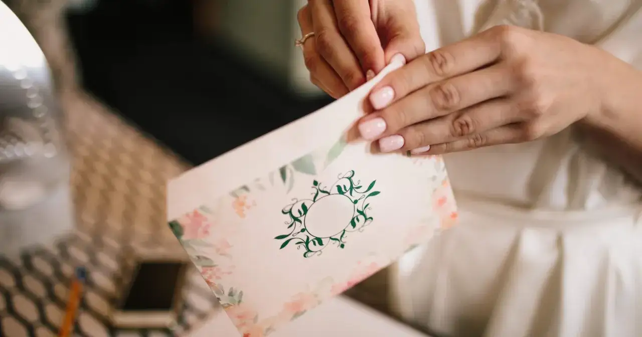 Dłonie z różowym manicure zamykają elegancką kopertę z kwiatowym wzorem. Jak sformułować zaproszenie na wesele bez dzieci, by nikt się nie obraził.