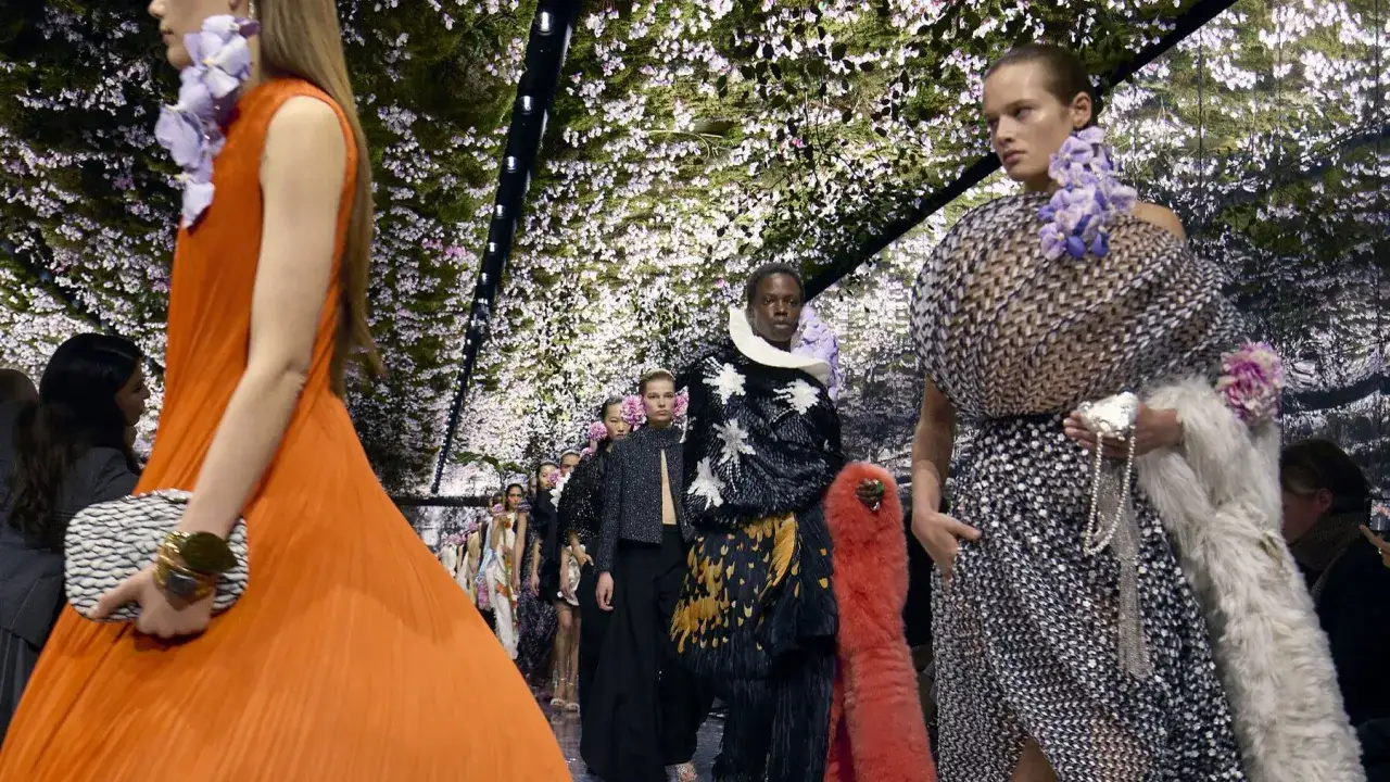 Sfilata Dior con modelli che indossano abiti floreali e pellicce, su uno sfondo di fiori bianchi e rosa. Una storia di moda.