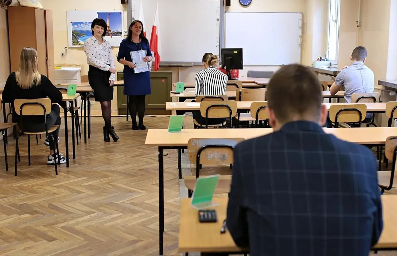 Uczniowie siedzą w ławkach, czekając na rozpoczęcie egzaminu zawodowego praktycznego. Nauczycielki tłumaczą zasady.