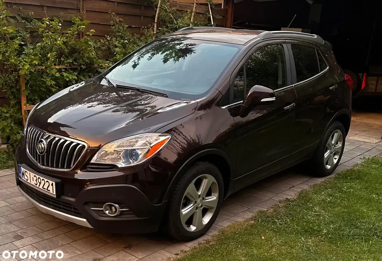 Brązowy Buick Encore zaparkowany na podjeździe z kostki brukowej, otoczony zielenią.