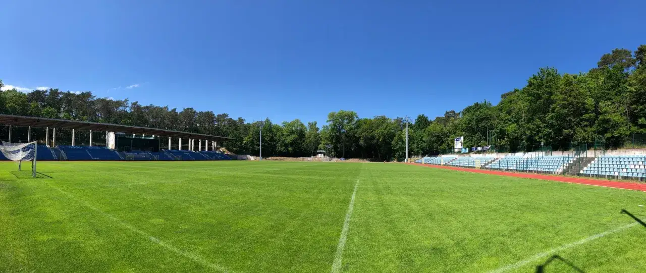 Zielona murawa i niebieskie trybuny stadionu Floty Świnoujście pod błękitnym niebem.