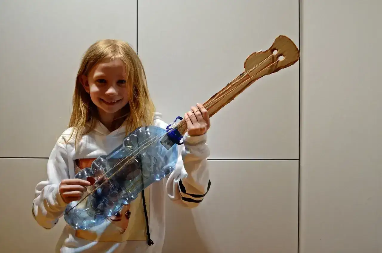 Dziewczynka z uśmiechem prezentuje własnoręcznie zrobiony instrument muzyczny do szkoły z butelki i kartonu.