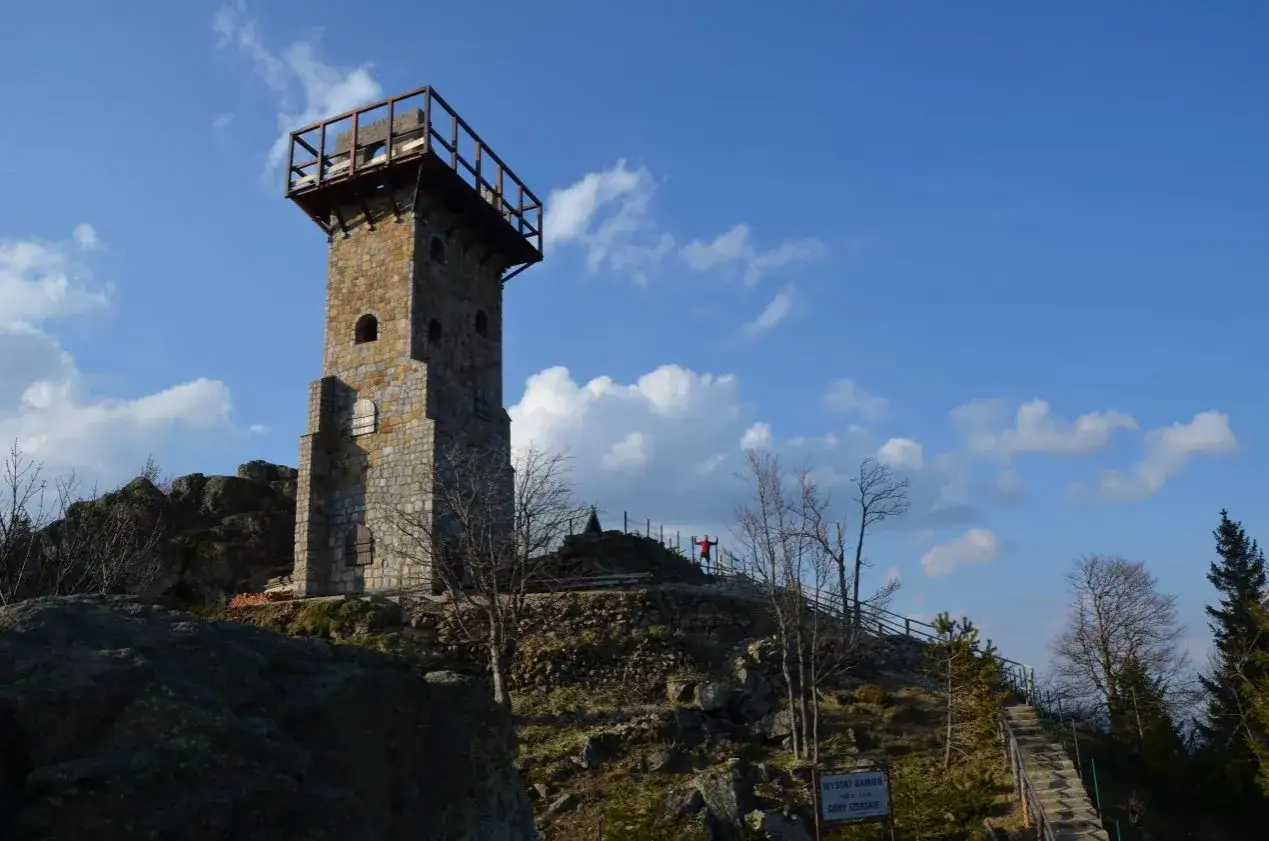 Kamienna wieża widokowa na szczycie wzg&oacute;rza, z drewnianym tarasem. W oddali osoba w czerwonej kurtce pozdrawia z platformy.