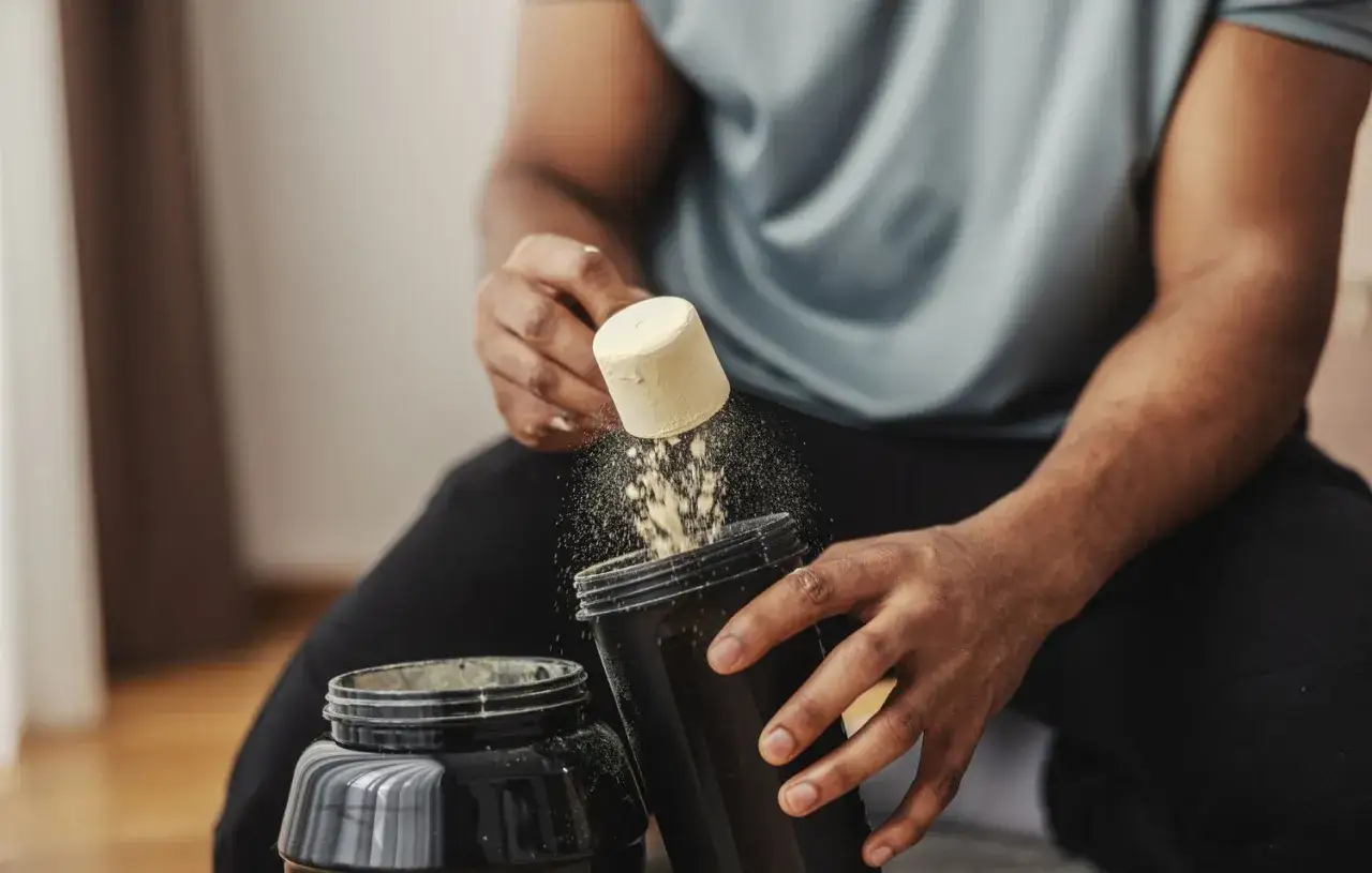 Mann bereitet einen proteinshake vor oder nach dem Training zu. Pulver rieselt aus einem L&ouml;ffel in einen Shaker.
