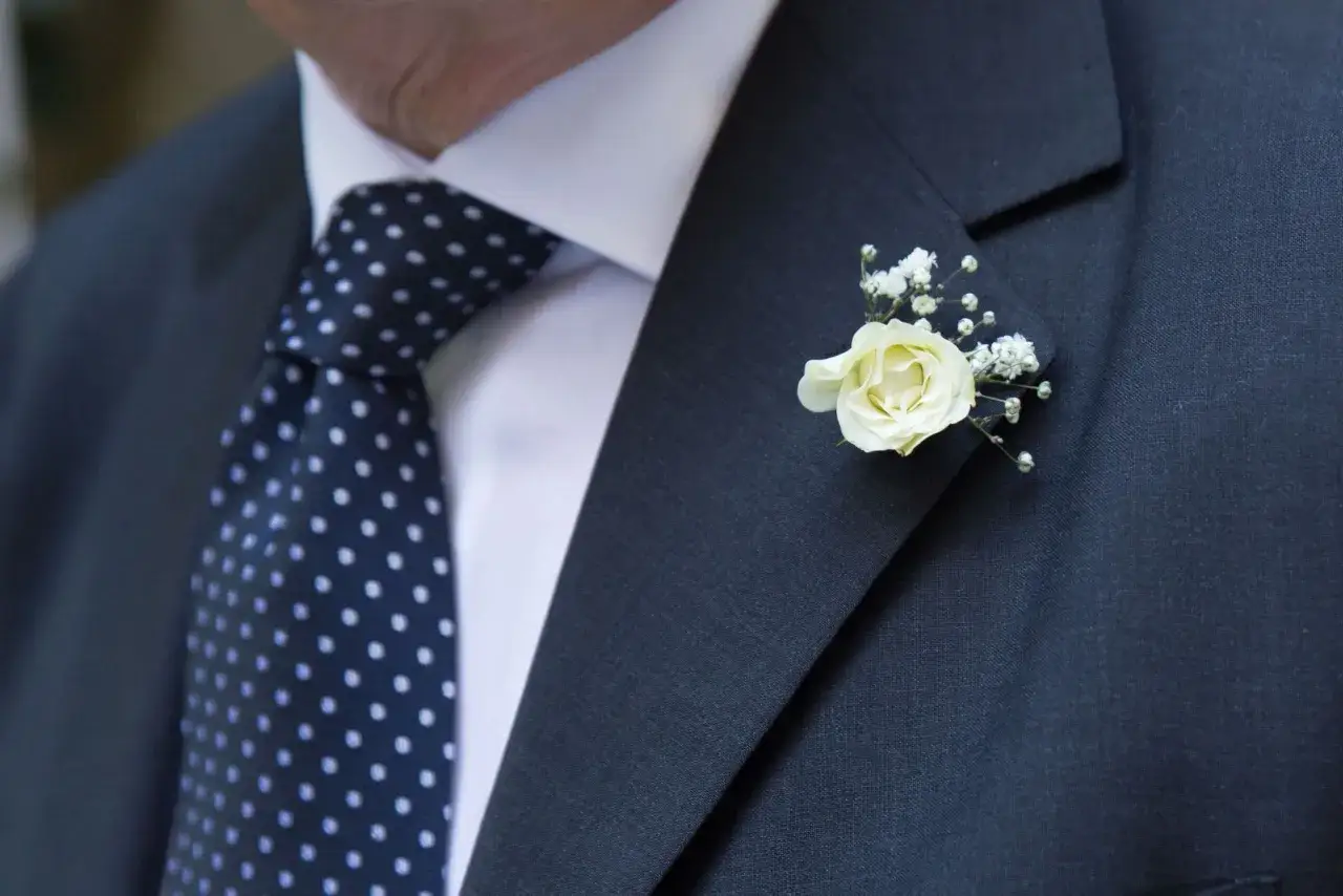 Elegancka butonierka co to? Delikatna biała róża z gipsówką zdobi ciemną marynarkę i granatowy krawat w kropki.