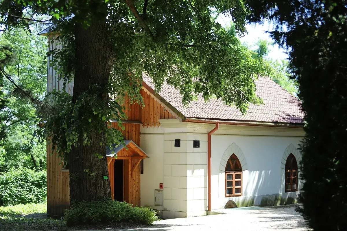 Drewniana dzwonnica i biały budynek z łukowymi oknami, otoczone zielenią. Idealne miejsce na jedne z atrakcji okolic Rzeszowa.
