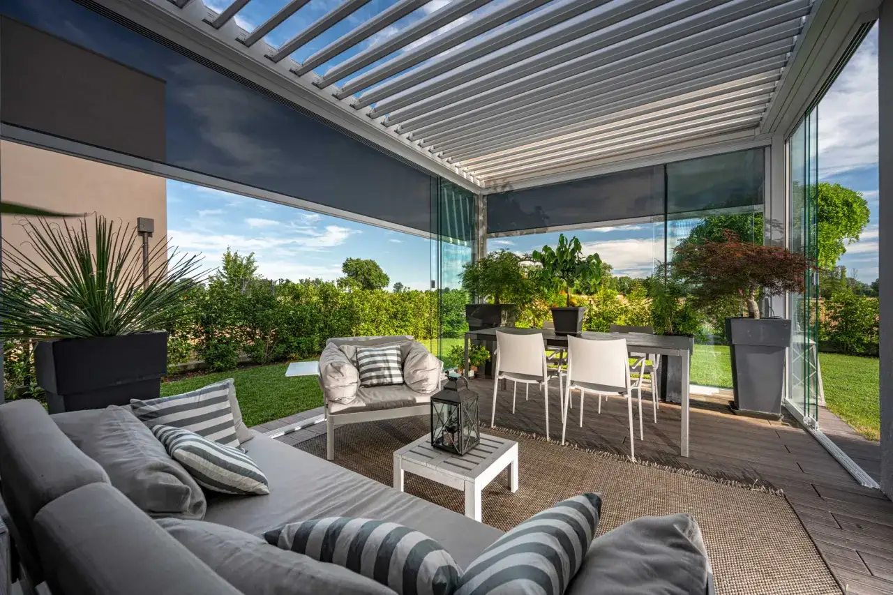 Una moderna pergola bioclimatica que es un espacio exterior con sof&aacute;s, mesa y sillas, rodeado de vegetaci&oacute;n.