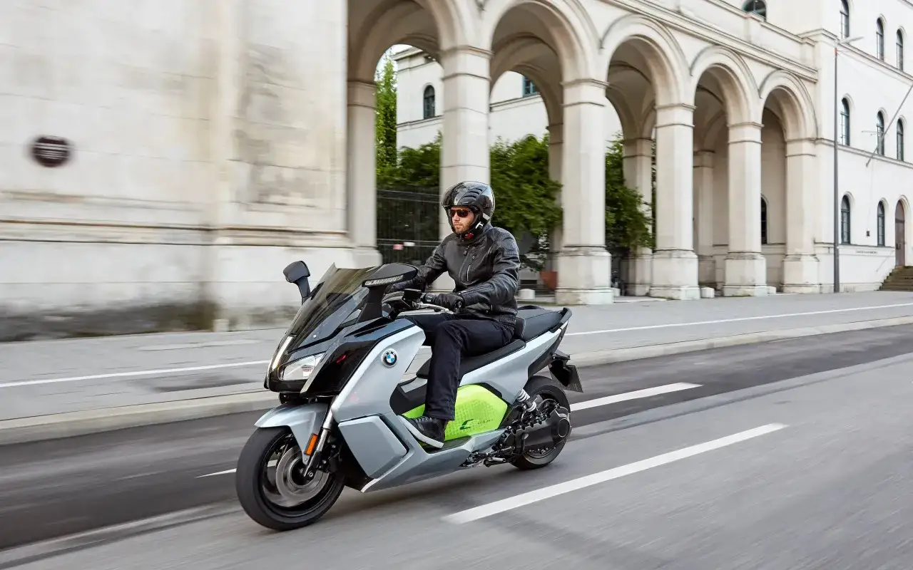 Mężczyzna w kasku i skórzanej kurtce jedzie nowoczesnym, srebrno-czarnym bmw skuterem elektrycznym z neonowo-zielonym akcentem, mijając zabytkową budowlę z arkadami.