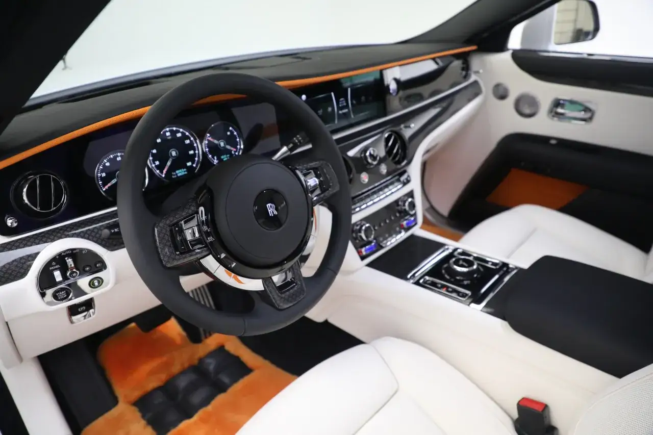 Wnętrze Rolls-Royce'a z białymi skórzanymi fotelami i pomarańczowymi dywanikami. Luksusowy kokpit z kierownicą i ekranami. Taki Rolls-Royce cena jest wysoka.
