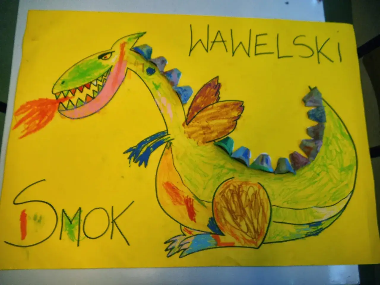 Kolorowa praca plastyczna przedstawia zielonego smoka wawelskiego ziejącego ogniem. Napis 