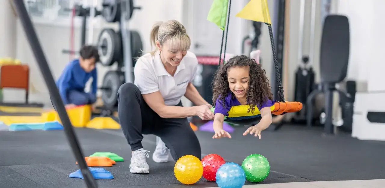 Therapeutin unterst&uuml;tzt ein Kind in einer Schaukel bei der Ergotherapie. Bunte B&auml;lle liegen auf dem Boden.