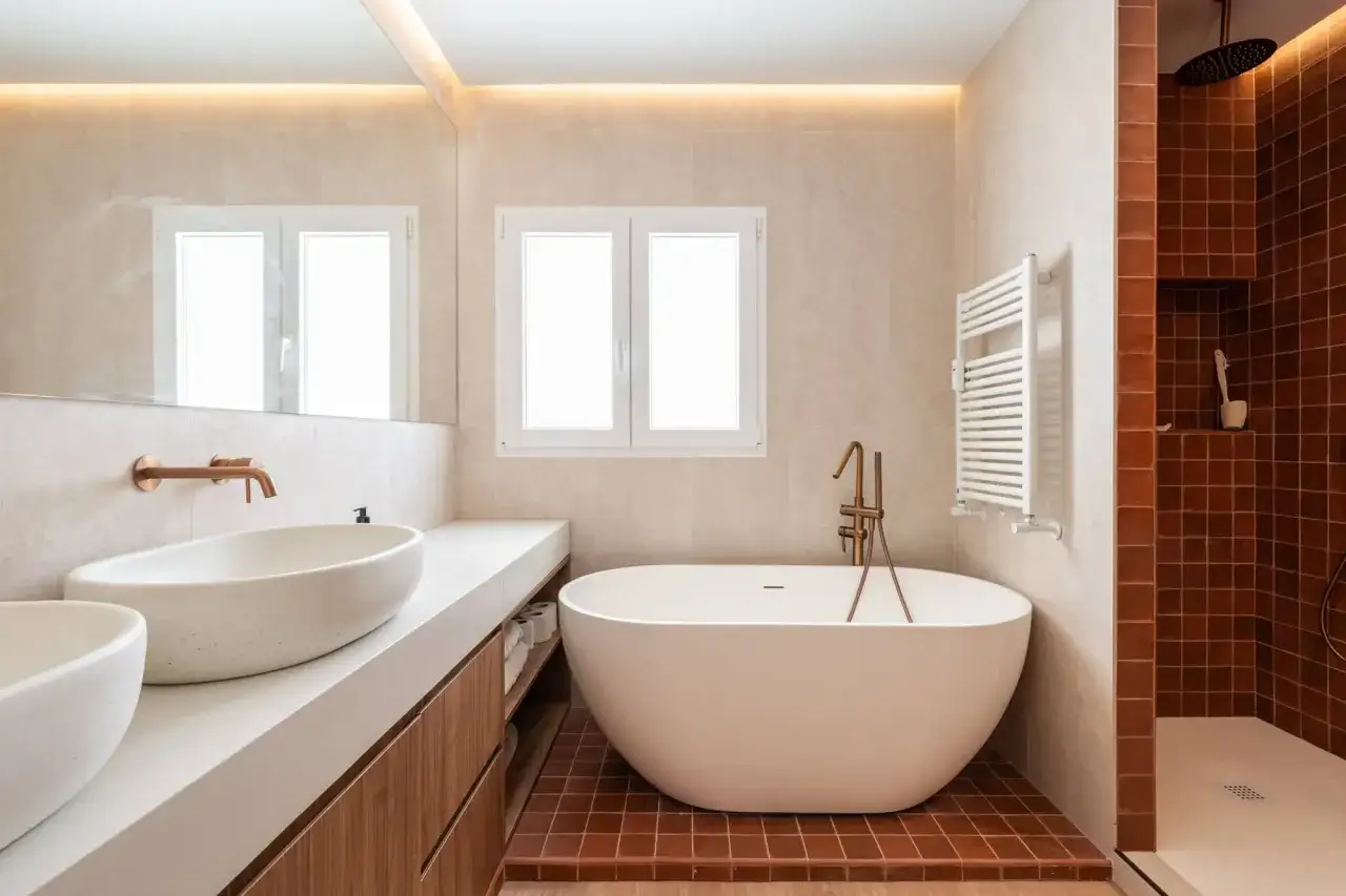 Ba&ntilde;o moderno con ba&ntilde;era exenta, lavabos sobre encimera y ducha. La iluminaci&oacute;n indirecta en el techo resalta la calidez de los materiales, una idea genial para saber como iluminar un ba&ntilde;o.