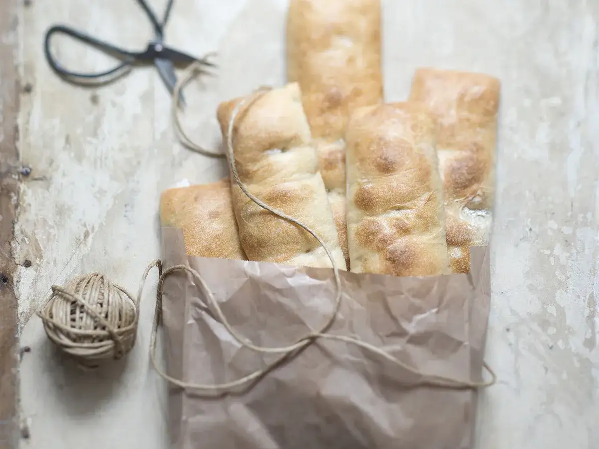 Un sac en papier brun contient plusieurs pains italiens dorés. Une ficelle et des ciseaux sont à proximité.