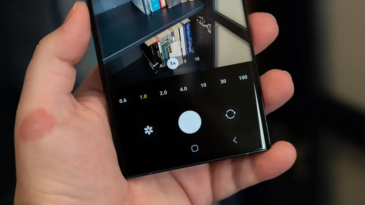 Nahaufnahme einer Hand, die ein Samsung-Handy h&auml;lt. Die Kamera-App ist ge&ouml;ffnet und zeigt eine B&uuml;cherregal-Szene. Man kann die Zoom-Optionen sehen, was das scannen mit handy samsung erleichtert.