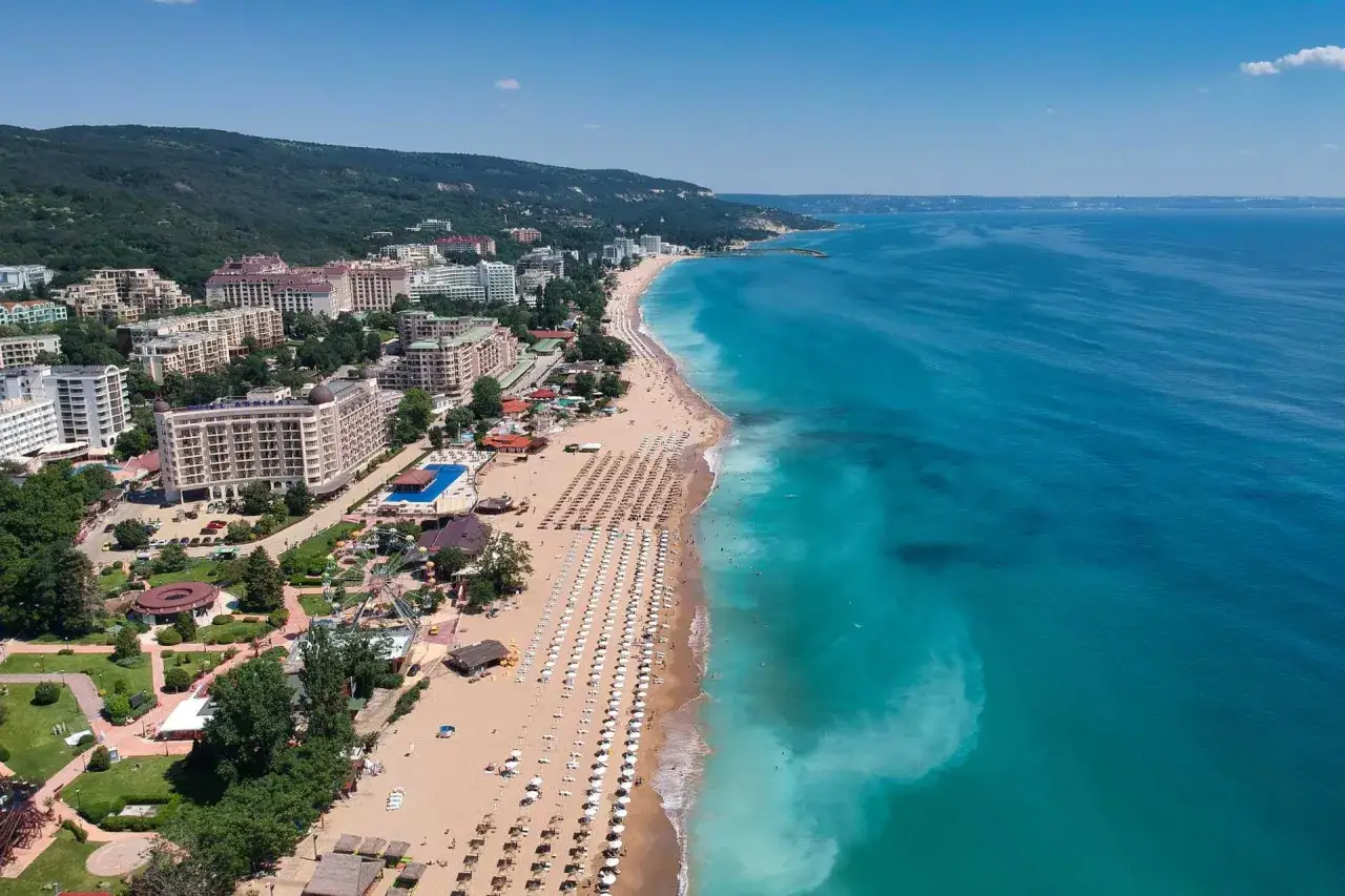 Widok z lotu ptaka na plażę w Złote Piaski, z hotelami, zielonymi terenami i diabelskim młynem. Atrakcje turystyczne w pełnym słońcu.