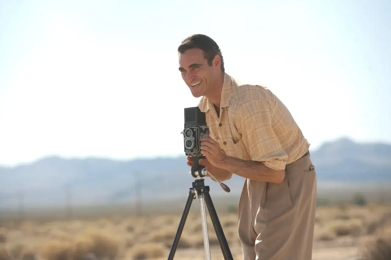 Hombre sonriente con c&aacute;mara Rolleiflex en tr&iacute;pode en un paisaje des&eacute;rtico, capturando la esencia de una pel&iacute;cula 70mm.
