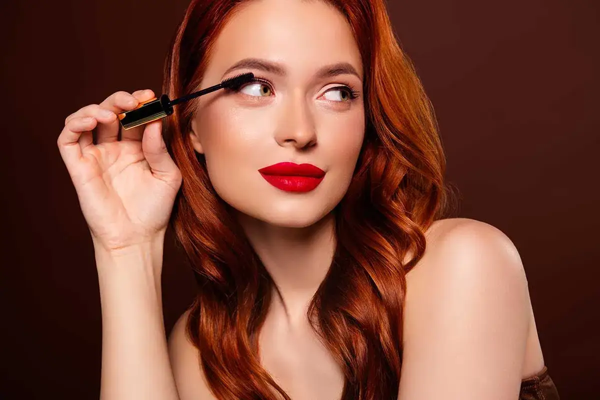 Donna con capelli rossi applica mascara, labbra rosse vivaci. Un look perfetto per sapere come truccarsi in estate.
