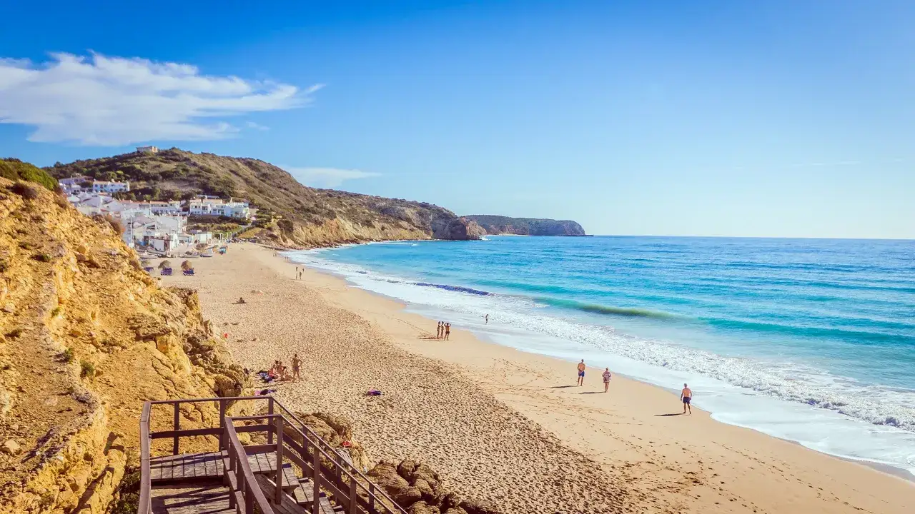 Sonne, Sand und Meer am Strand von Salema, Portugal. Weiße Häuser schmiegen sich an grüne Hügel, während Menschen am Ufer spazieren.