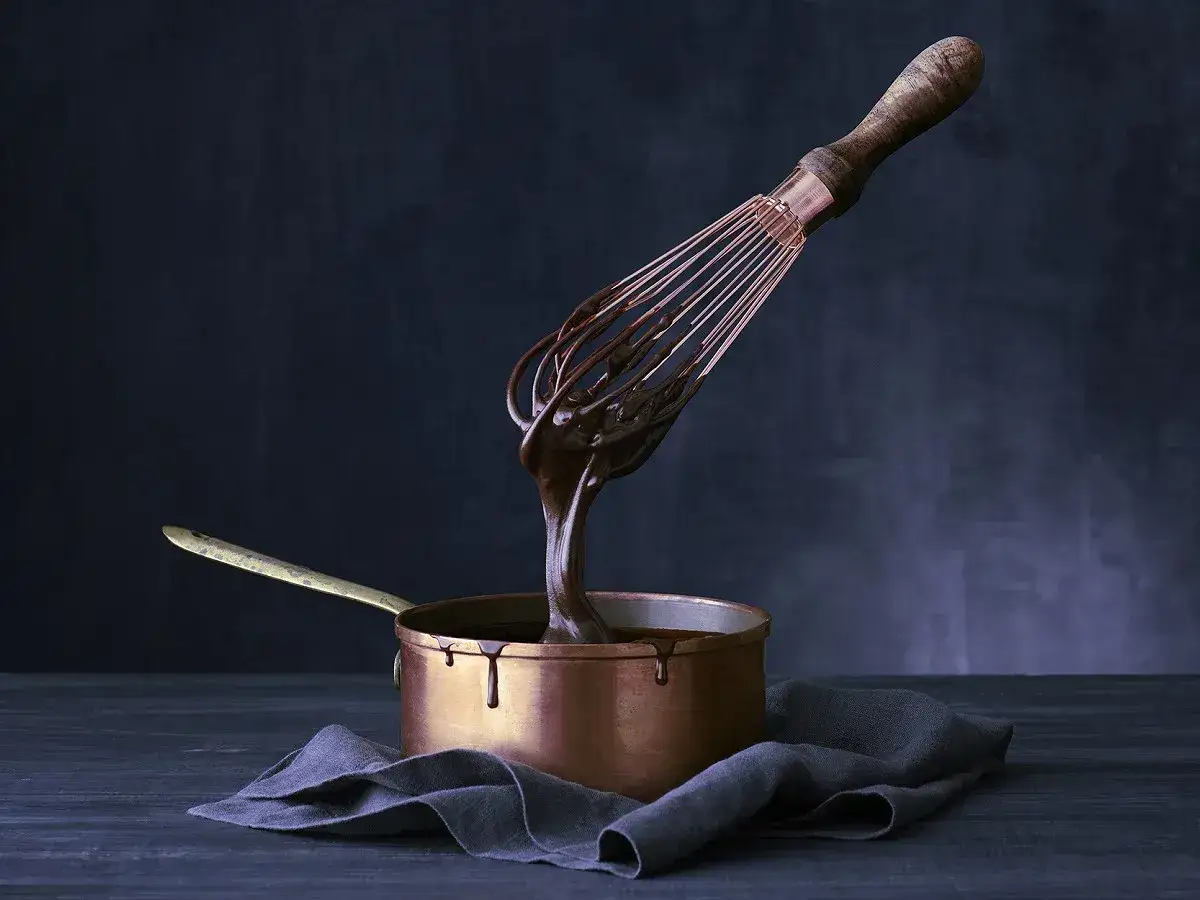 Un fouet en cuivre dégouline d'une sauce chocolat onctueuse dans une casserole en cuivre.