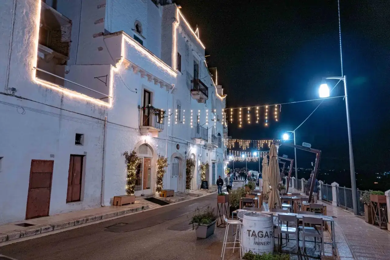 Un incantevole **locorotondo di sera**, con luci natalizie che illuminano i bianchi vicoli e creano un'atmosfera magica. Tavolini all'aperto invitano a godersi la quiete notturna.
