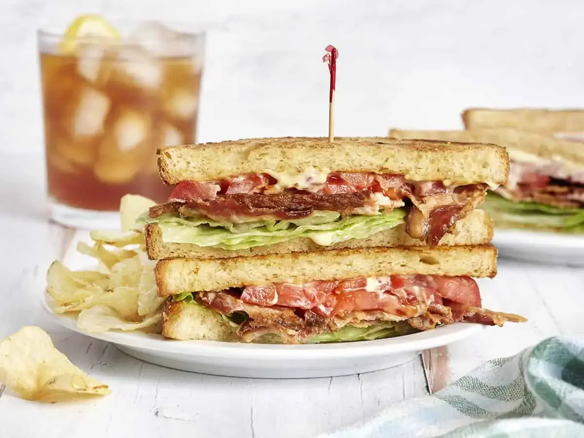 Ein üppiger BLT-Sandwich-Turm mit knusprigem Speck, saftigen Tomaten und knackigem Salat, serviert mit Kartoffelchips und Eistee.