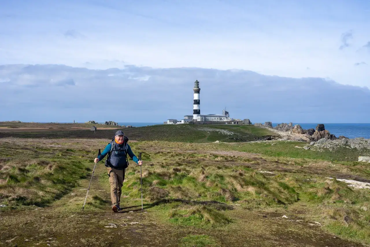 Un randonneur souriant avec des bâtons de marche explore Ouessant à pied, le phare emblématique en arrière-plan.