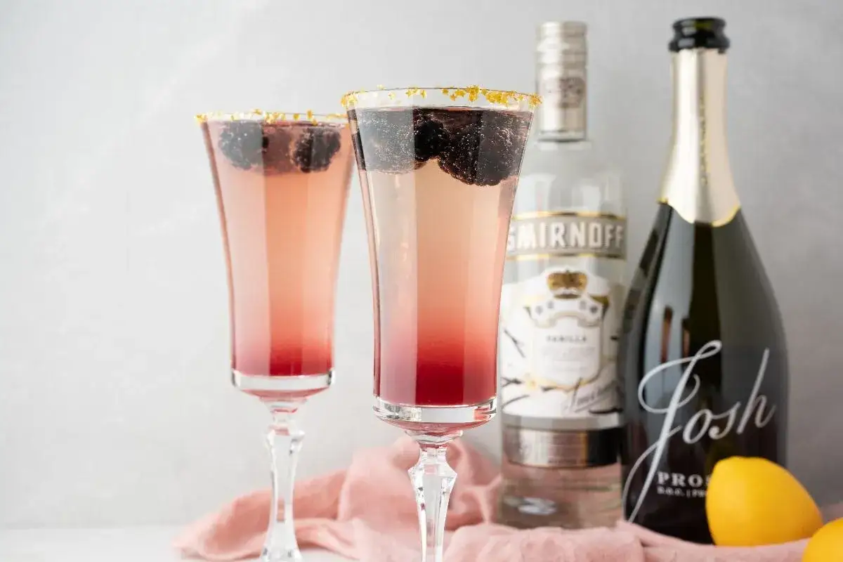 Dwa kieliszki z różowym prosecco drinkiem, ozdobione jeżynami i skórką cytrynową. Obok butelka wódki i prosecco.