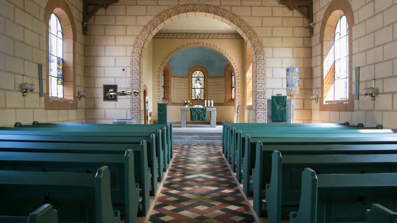 Leere Kirchenbänke in Grün laden dazu ein, dass man wieder in die Kirche eintreten kann. Buntglasfenster und ein Altar im Hintergrund.