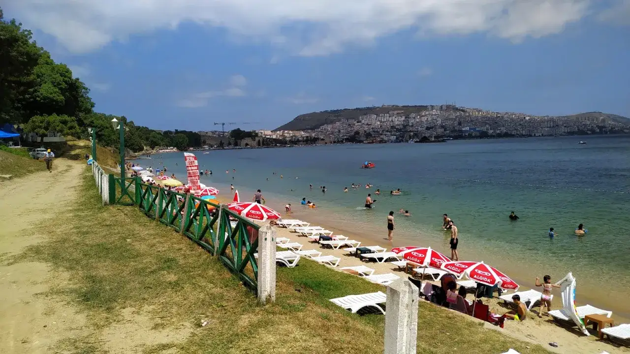 Słoneczny dzień na plaży w **Sinop**. Ludzie kąpią się w morzu, opalają na leżakach pod parasolami. W tle widać miasto na wzgórzu.
