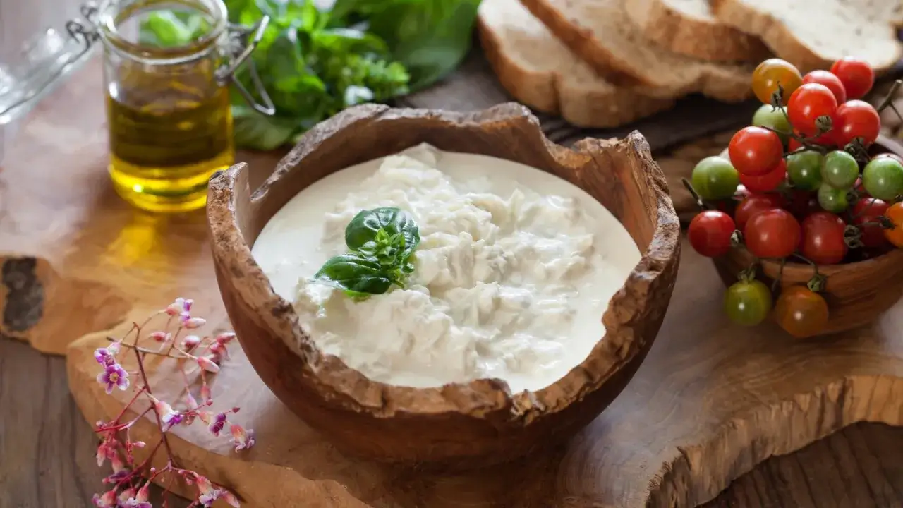 Kremowa stracciatella w drewnianej misie, ozdobiona bazylią, obok pomidorków koktajlowych i oliwy.