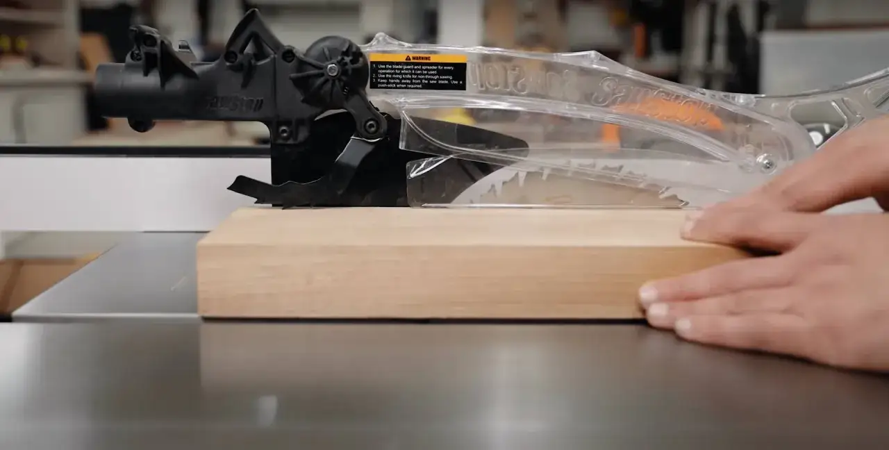 Hands guide wood through a table saw blade, demonstrating how to use a table saw safely with the blade guard in place.