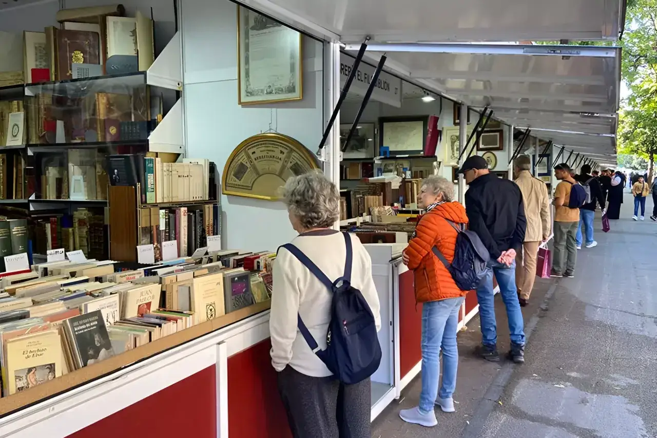 Gente buscando tesoros literarios en la feria del libro antiguo de Madrid. Estanter&iacute;as repletas de tomos esperan ser descubiertos.