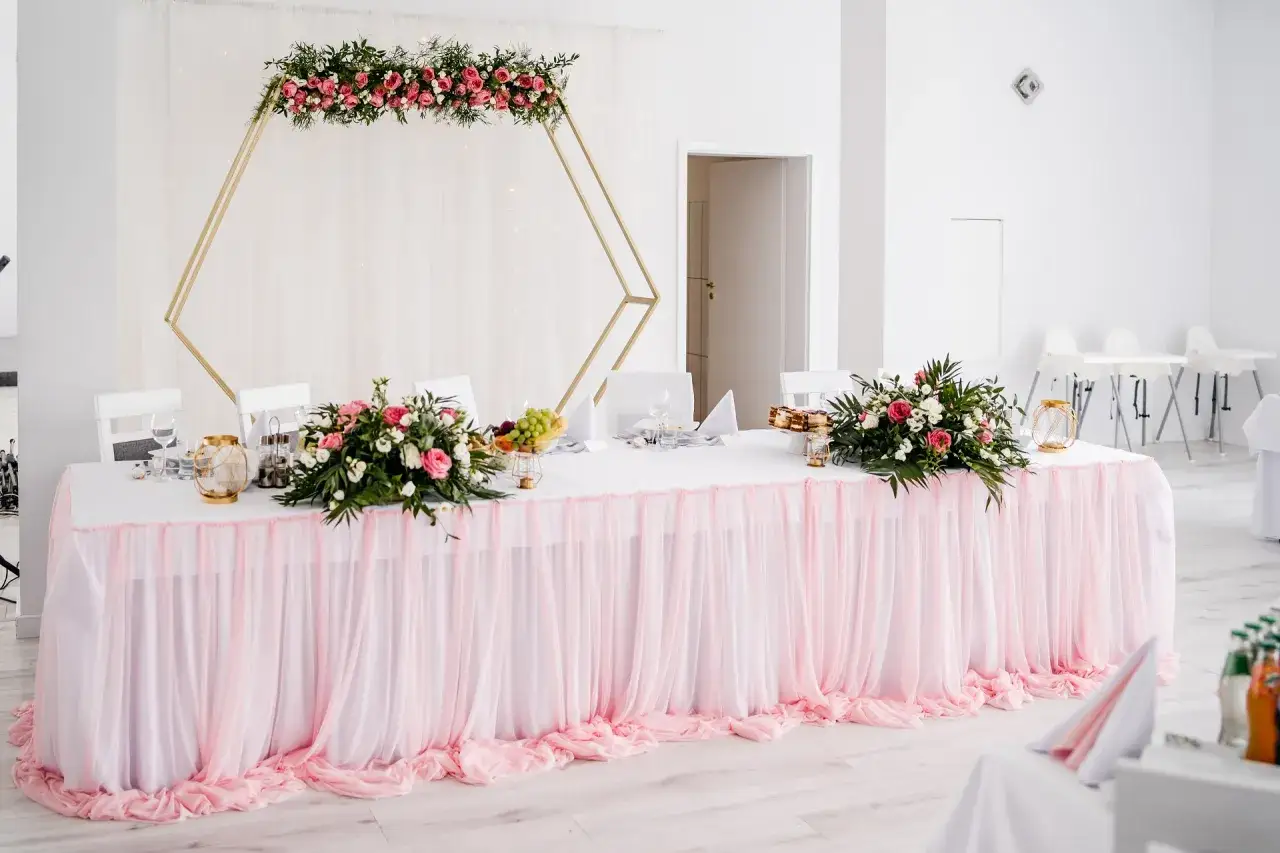 Elegancka dekoracja sali weselnej: złota konstrukcja z kwiatami, różowy tiulowy obrus i bukiety róż. Inspiracje na przyjęcie.