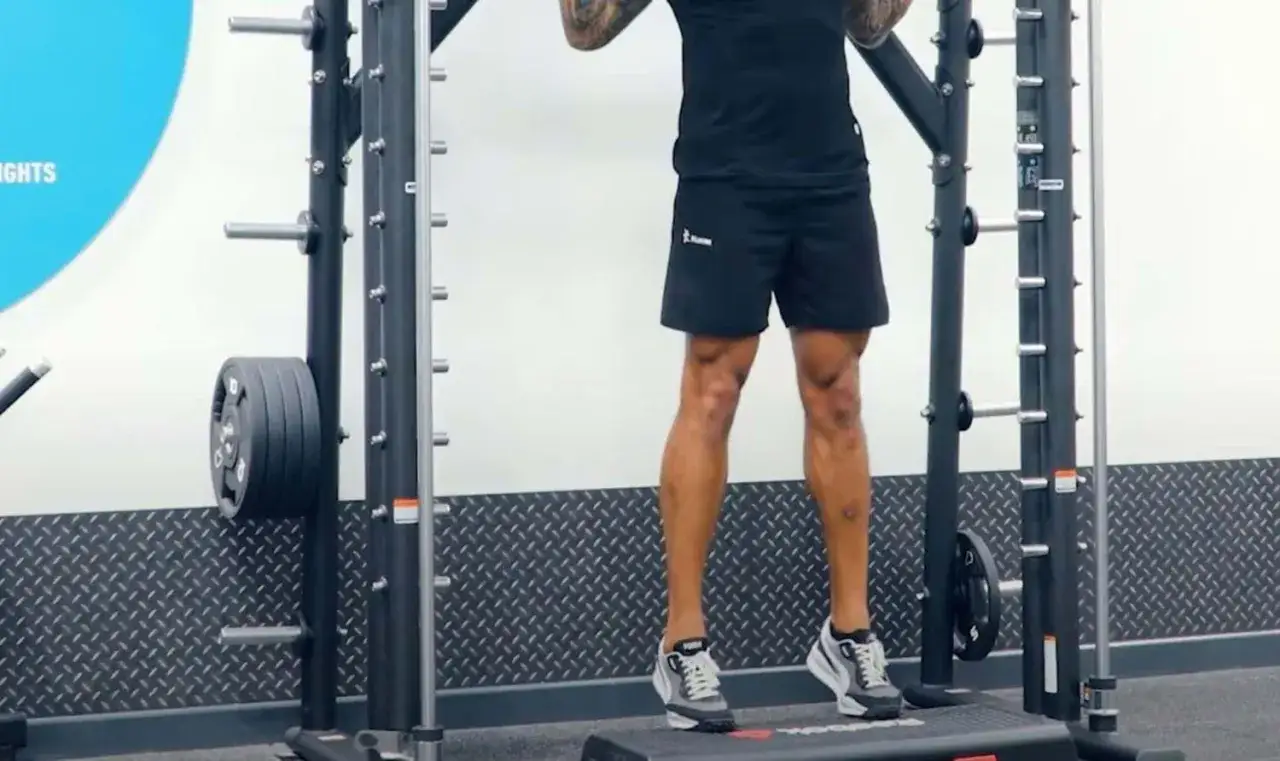 Uomo in palestra esegue calf raise su step, con bilanciere appoggiato sulle spalle.