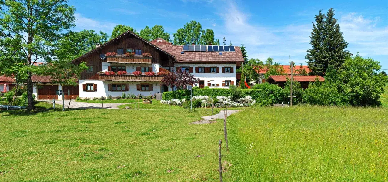 Ein idyllischer Bauernhof in den Bergen, umgeben von grünen Wiesen und Bäumen. Das Haus hat traditionelle Holzdetails und Solarpanels auf dem Dach.