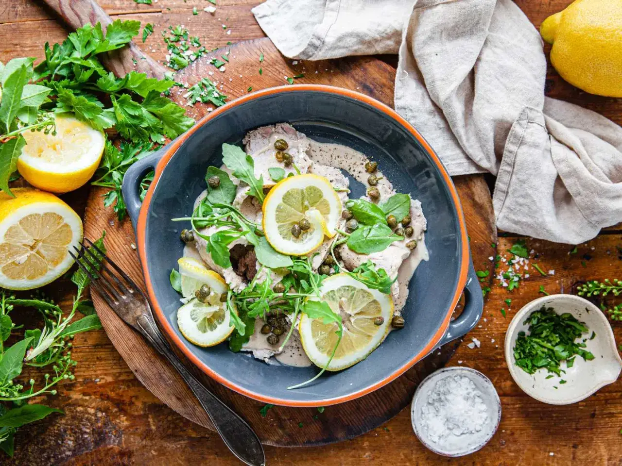 Vitello tonnato i en gr&aring; sk&aring;l, toppad med citronskivor, kapris och ruccola. Serveras med f&auml;rska &ouml;rter och citroner p&aring; ett tr&auml;bord.
