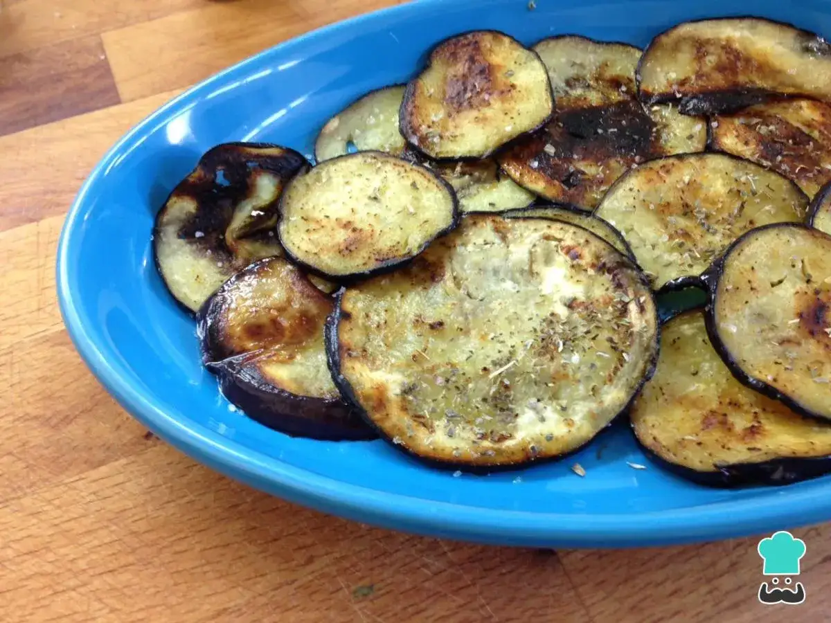 Berenjenas a la plancha, doradas y sazonadas con hierbas, servidas en un plato azul sobre una tabla de madera.