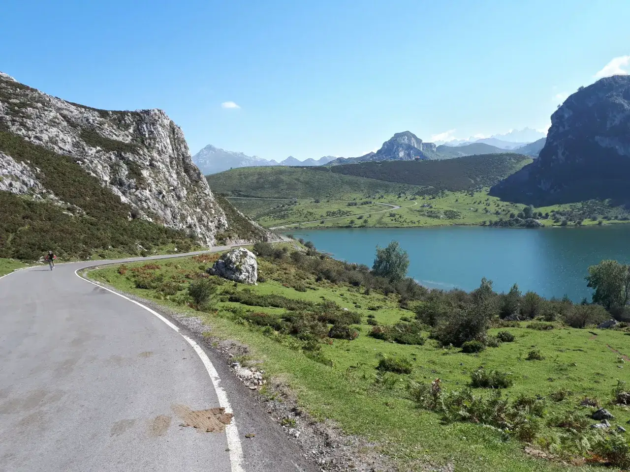 Ciclista en carretera serpenteante con lago turquesa y monta&ntilde;as de Picos de Europa cerca de Llanes.