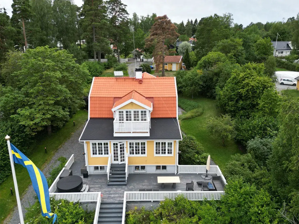 Gult hus med orange tak och en takkupa, omgivet av grönska. En altan med möbler och en svensk flagga. En utbyggnad av taket har gjorts.