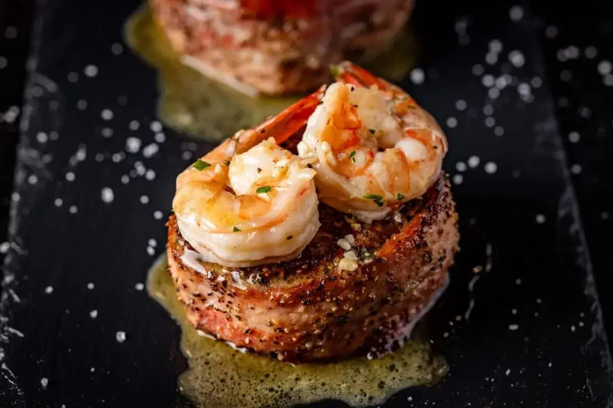 Ein k&ouml;stliches Surf and Turf Rezept: saftiges Steak umwickelt mit Speck, gekr&ouml;nt von zwei Garnelen, serviert auf einem schwarzen Teller mit zersto&szlig;enem Salz.