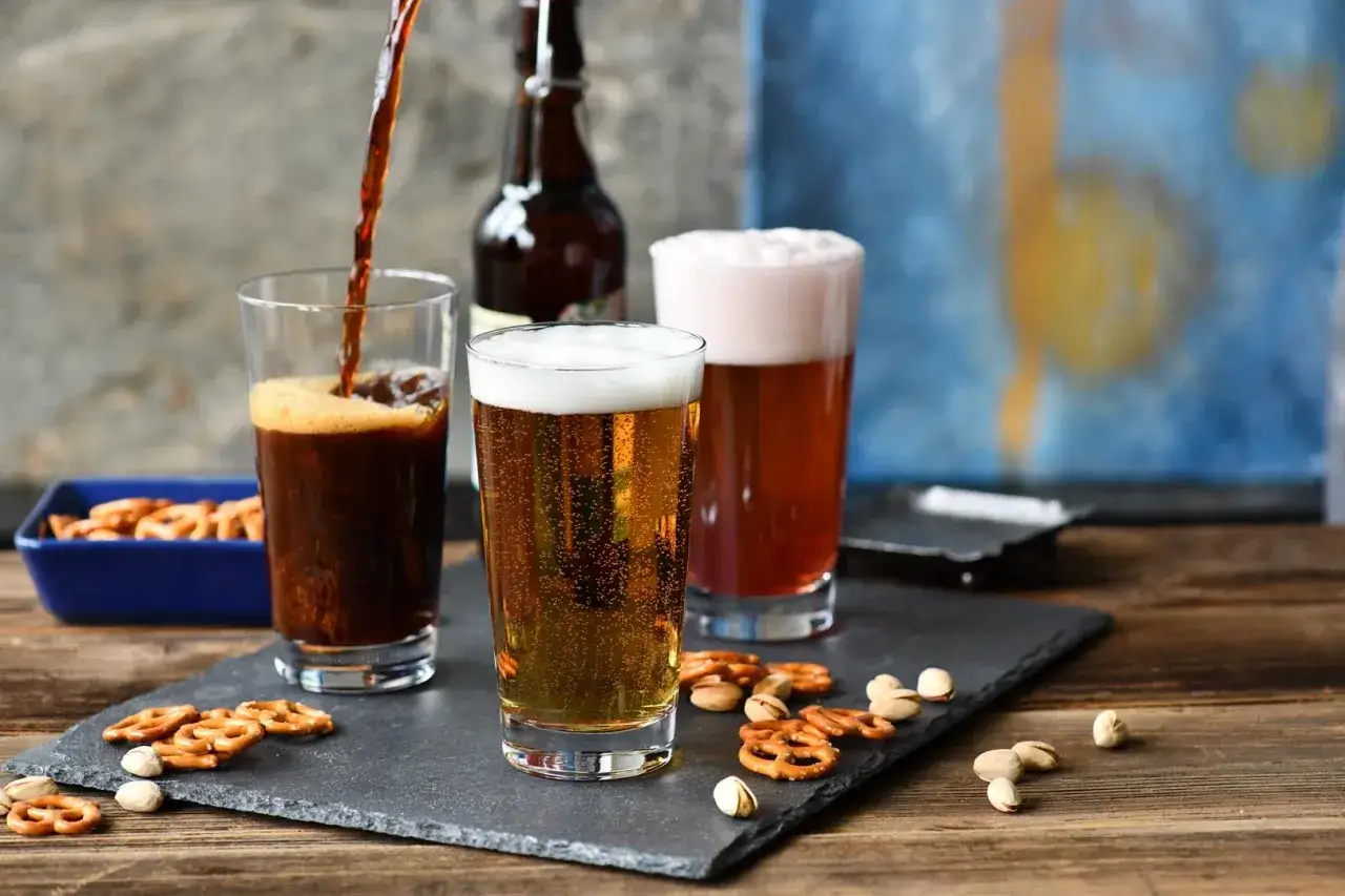 Jak zrobić domowy radler z piwa jasnego i lemoniady? Trzy szklanki piwa, precelki i pistacje na kamiennej desce.