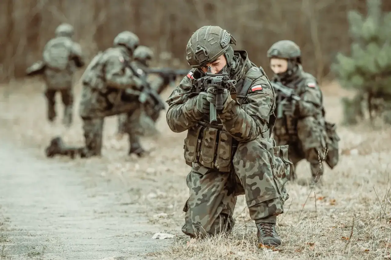 Szeregowy w wojsku w pozycji klęczącej celuje z karabinu, podczas gdy inni żołnierze zajmują pozycje w tle.