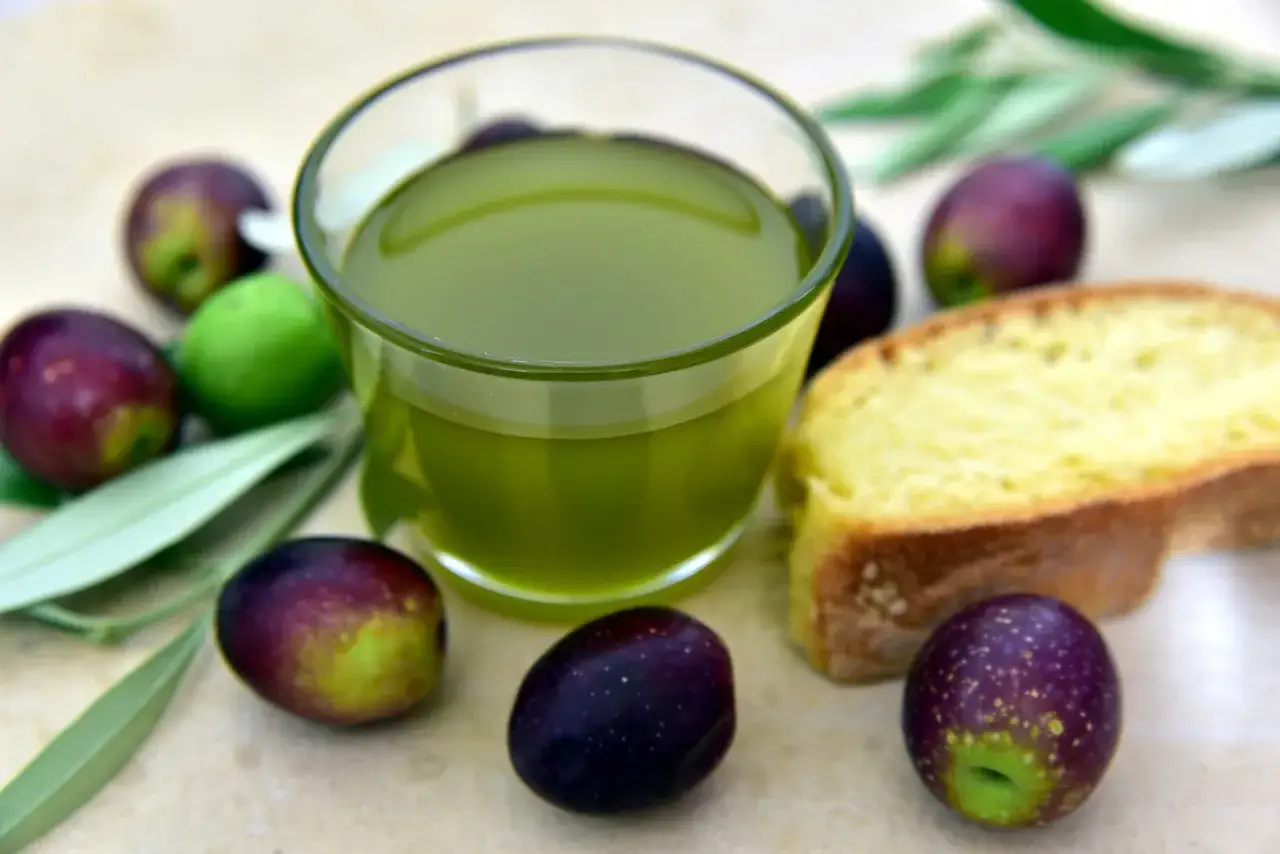 Un bicchiere di olio d'oliva verde brillante, olive fresche e una fetta di pane. Si può mischiare l'olio d'oliva con l'olio di semi per un condimento versatile.