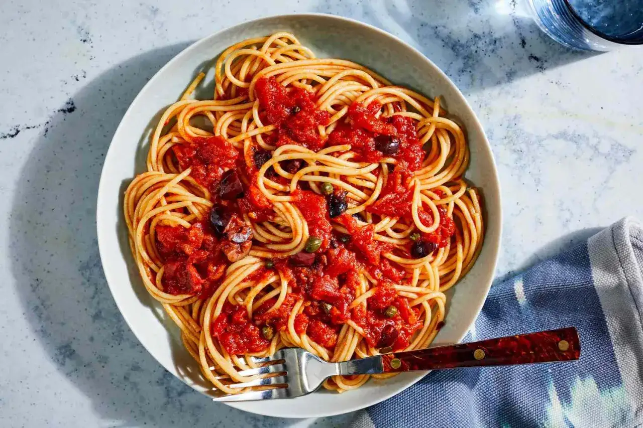 Eine Schüssel Spaghetti Puttanesca mit Oliven, Kapern und Tomatensauce, serviert mit einer Gabel.